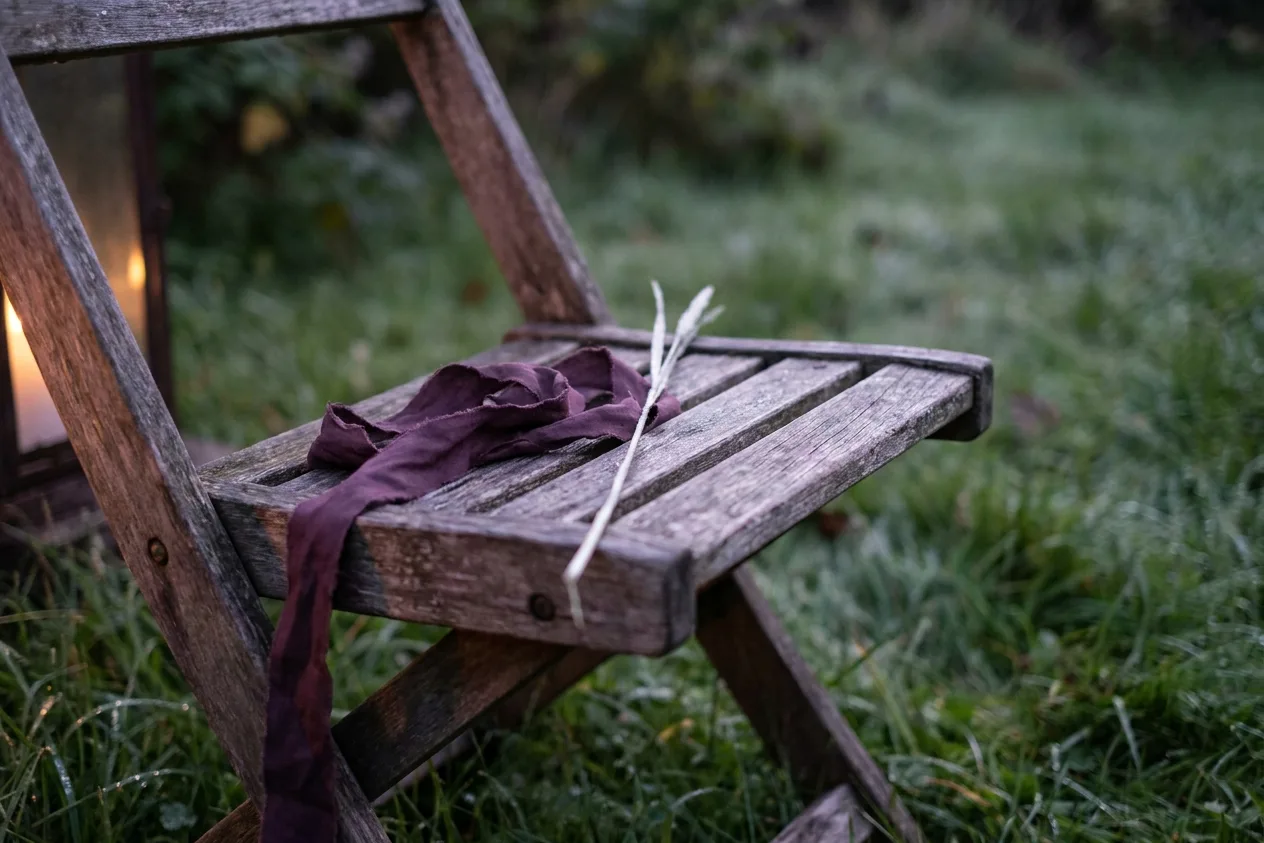 A folded plum ribbon resting on the slats of a weathered wooden folding chair on frosted grass, a lantern glowing behind it.